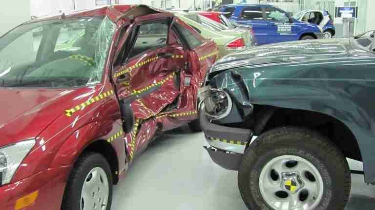 Side-impact collision demonstration showing significant damage to a red car with crumple zones indicated by yellow markings.